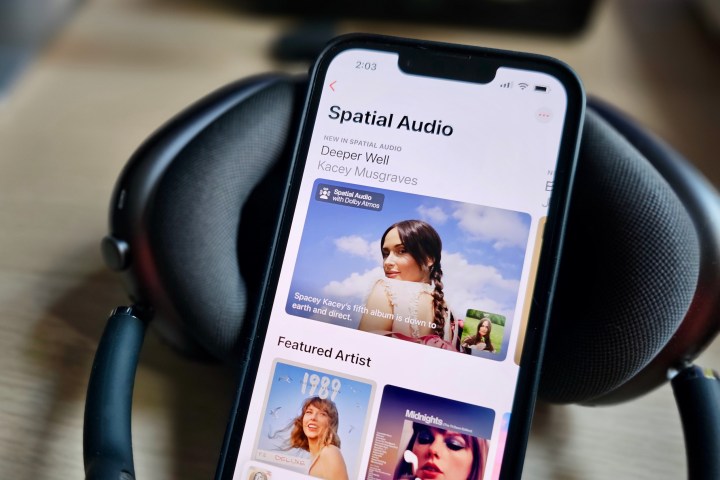 هاتف Apple iPhone 14 يعرض تطبيق Apple Music مع صفحة ميزات حول محتوى الصوت المكاني، بجانب مجموعة سماعات Apple AirPods Max.